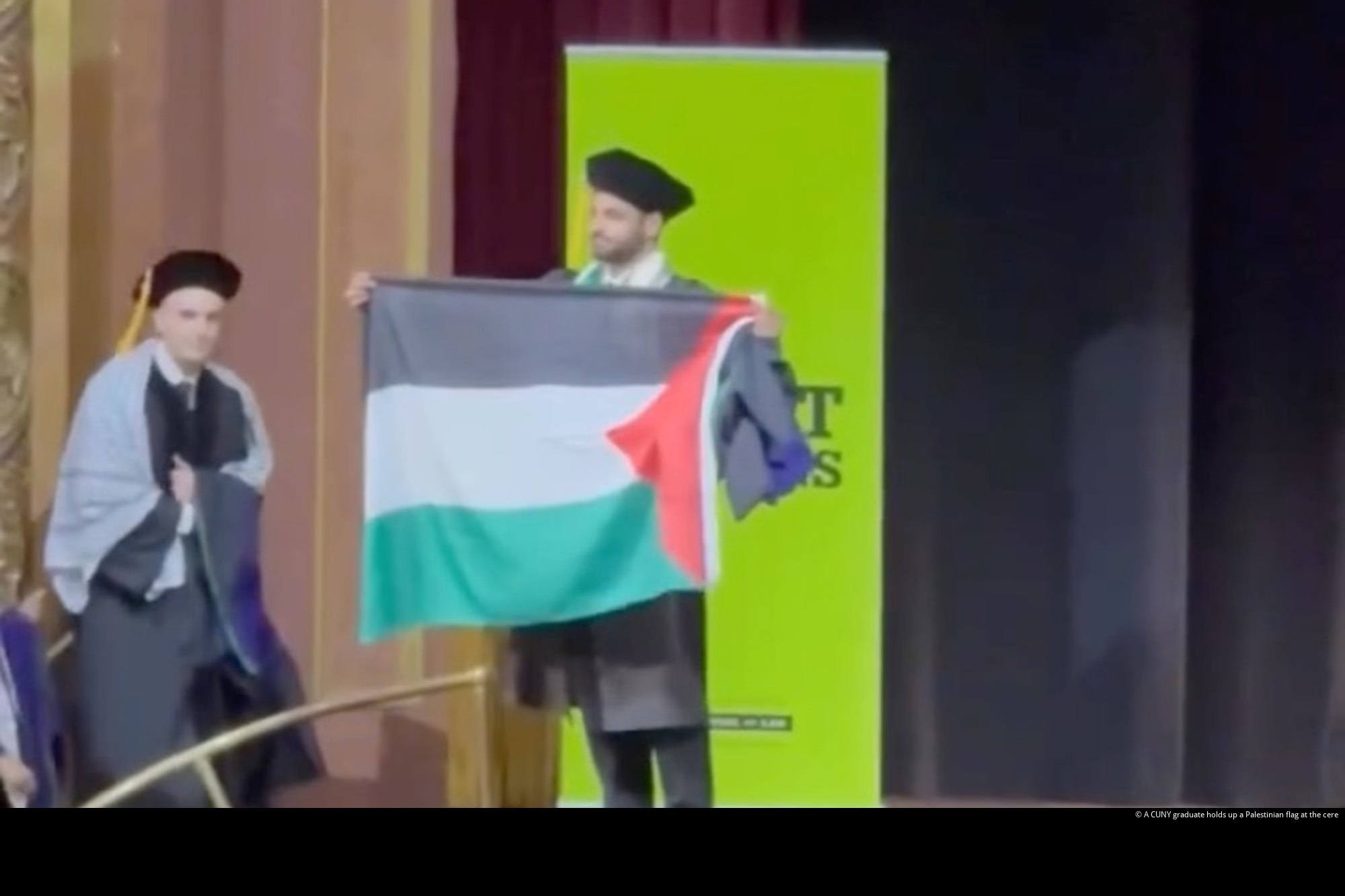 A graduate in cap and gown holds up a Palestinian flag on stage.